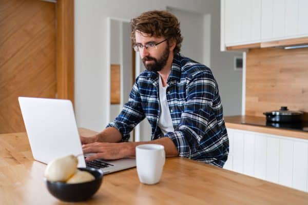 Man with beard  in  plaid shirt  using  laptop in  the  kitchen.
